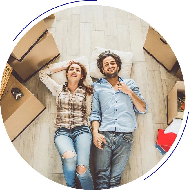 A man and woman lie on the floor amidst packing boxes, smiling and holding hands.