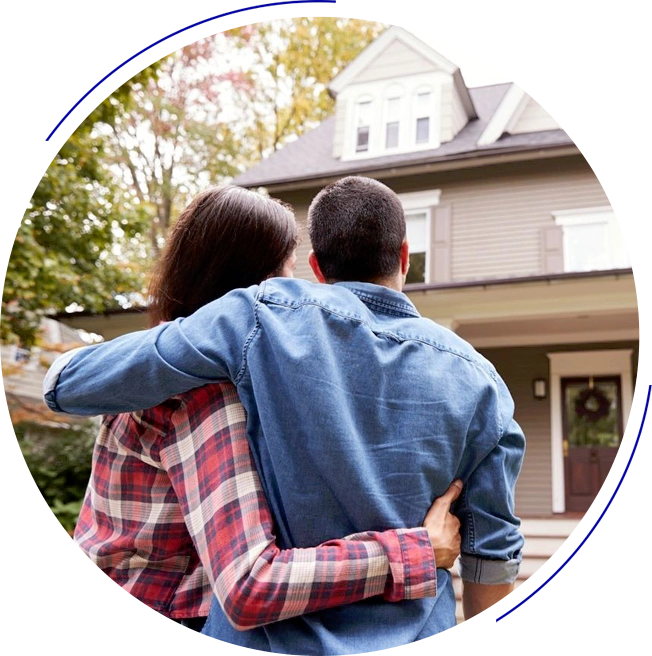 A couple stands with their backs to the camera, arm in arm, looking at a two-story house with a front porch. The house is surrounded by trees and has a wreath on the front door.