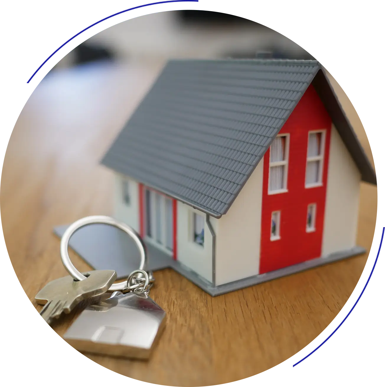 A small model house with red and white exterior is placed on a wooden surface. Two keys on a keyring rest beside it.